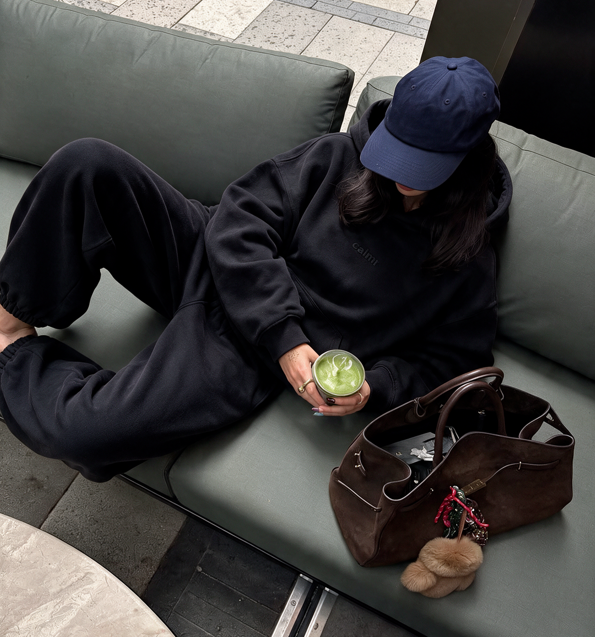 A person wearing a black hoodie and black sweatpants lounges on a green sofa, holding a green drink. A brown leather tote bag and a small beige bag rest beside them on the couch.