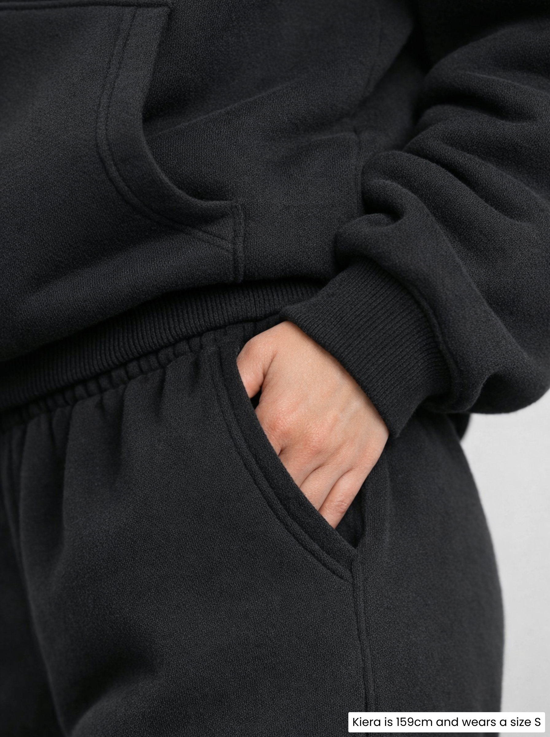 Person wearing a slightly weighted black hoodie and pants with one hand in the pocket against a light background