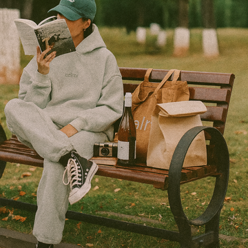 Person sitting on a park bench reading a book with bags and a bottle beside them.